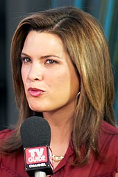 An image of a woman with shoulder-length hair and a dark red outfit. She is speaking into a microphone with the words "TV Guide".