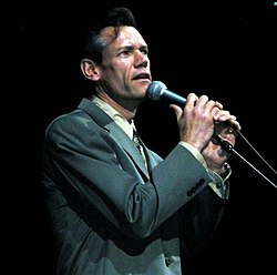 A dark-haired man in a grey sports coat singing into a microphone