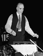 elderly, balding man with short white moustache and beard, conducting an orchestra