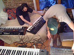 Musician with headphones playing a synthesizer, while another musician adjusts an equalizer