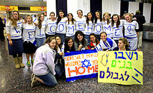 Photograph at an airport of a welcoming group for new immigrants from North America. Several girls are kneeling in the foreground holding signs with welcoming messages in both English and Hebrew while 11 others are standing behind them with the individual letters of the phrase "Welcome home!" drawn in blue on white shirts.