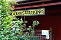Ein gelbes Schild mit dem Namen „Werstattkino“ hängt über einem verschnörkelten Geländer, von wo aus es die Treppe hinunter in den Keller zum Kinoraum geht