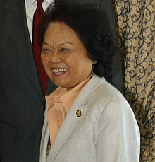 Photograph of an Asian woman in an orange blouse and white blazer