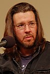 David Foster Wallace is pictured with shoulder-length hair and a short beard. Wearing frameless glasses, he is speaking at a microphone and is wearing a black denim jacket over a t-shirt.