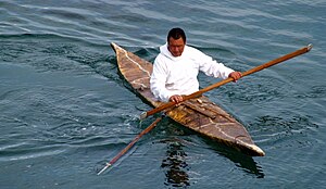 Man sitting with legs covered in a boat that tapers to a point at each end holding long, pointed, wooden pole
