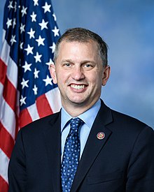 Official portrait of Casten from the 117th Congress. Sitting in front of an American flag, he wears a dark suit, a light blue shirt, and a blue tie.