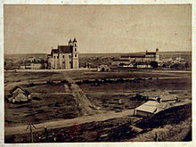 Schwarzweißfotografie in der Obersicht von zwei großen Kirchenbauten und mehreren Häusern im Hintergrund. Im Vordergrund sind Felder, Wege und einige Hütten