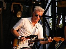 A man with gray hair, wearing a white shirt and sunglasses, plays an electric guitar.