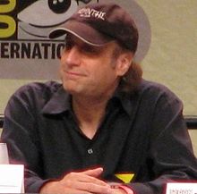 A seated David Mirkin wearing a cap smiles as he looks into the distance. His hands are crossed.
