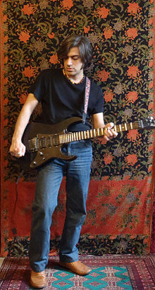 Salim Ghazi Saeedi wearing blue jeans and a dark t-shirt, standing on a patterned rug with a patterned drape as a background, playing a black electric guitar
