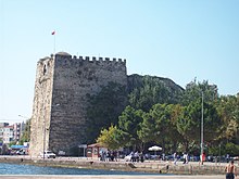 Photograph of a tall, roughly square stone fortress in a modern coastal city.