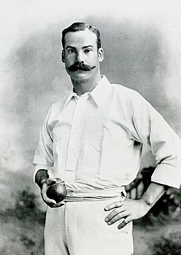 Mold standing wearing cricket whites and holding a cricket ball in his right hand