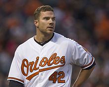 A Caucasian man with short-cropped light brown hair and hazel eyes, shown from about the waist up, looks to the right. He is wearing a short-sleeved white button-front baseball jersey, with "Orioles" written in stylized orange script trimmed in black across the front, above the similarly treated number 42, over a black T-shirt with the Nike swoosh logo visible at the neck. Behind him is an indistinct, blurred crowd