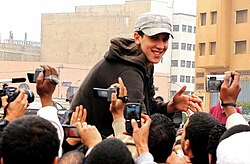 Belaghouat wearing a dark hoodie and light cap, being filmed by a crowd of people, smiling and appearing to shake hands