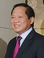 Trương Minh Tuấn seen smiling, wearing a black suit, a red tie and a blue shirt.
