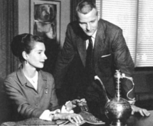 A white woman seated at a desk and a white man leaning over the desk; both are wearing suits, and gazing at a Yemeni vessel resembling a teapot
