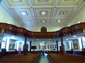 The interior looking towards an arched exit. There are rows of pews and a wide centre aisle. The ceiling is coffered into decorative square shapes. There is a large gallery of dark wood with a curving balustrade across the back of the church.