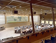 A large room with console arrayed in front of a world map and many desks occupied by people viewing screens