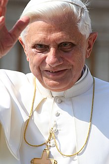 Farbige Nahaufnahme vom winkenden Benedikt 16 in weißem Papstgewand mit goldenem Kreuz als Halskette.