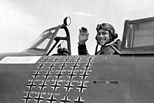 A fighter pilot sitting in an aircraft cockpit, shown in profile, viewed from the left. The pilot is smiling and waving his right hand in the air. The left side of the cockpit bears approximately 25 small black crosses arranged in five rows and five columns.