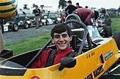 A man in his early twenties sitting in a stationary yellow racing car