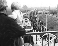 Mann mit Kleinkind auf dem Arm steht auf einer Brücke, die Kamera sieht an ihm vorbei auf einen Zug an einem Bahnsteig, dort steigen viele Arbeiter aus.