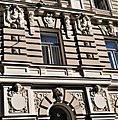 Farbige Nahaufnahme eines Fassadenteils mit Kopfreliefs zwischen den Fenstern und einem Tierkopf über dem Torbogen.
