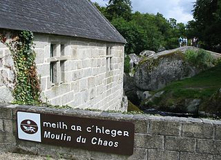 Side of a stone building next to a stream; low stone wall in the foreground has a sign reading Mill of Chaos in both Breton and French; Meilh ar Cʼhlegr and Moulin du Chaos