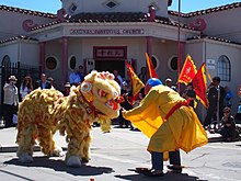 A costume lion and man in garb play fight at the Salinas Asian Festival, in front of the red and mauve confucius church
