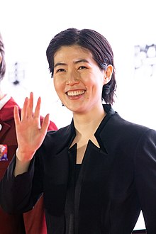Shim at press event smiling and waving one hand toward the camera