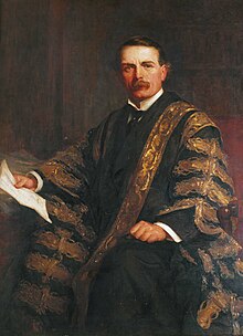 A man in his late 40s, with brown hair and moustache, wearing a black suit, white wing-collared shirt and black tie, together with a black gown repeatedly patterned in gold brocade; he sits in a chair, holding a piece of paper in his right hand