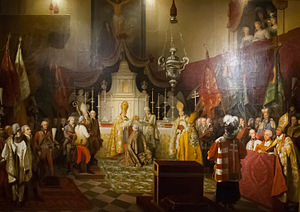 Interiro of a Catholic church. Before the altar, kneels a man in a golden robe. A bishop in gilded clothesand a layman in white are holding the Crown of Saint Stephen above his head. Around them stand many bishops and laymen in courtly clothes.