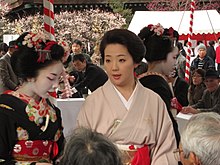 A geisha wearing a plain pink kimono with no white face makeup stood to the right of a maiko in full makeup wearing a heavily decorated black kimono.