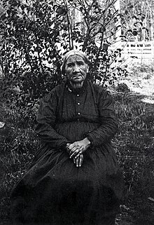 A photograph of an elderly woman with her hands in her lap, wearing a patterned dress, and sitting in front of a bush.