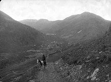A man on a horse wearing a jacket and hat, looking out into valley from the Otto Mears road; road leads down to the town of wood-frame structures and the Gold Prince Mill. Photo circa 1888.