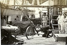 A man practices aiming a small gun on a wheeled, high-angle carriage while three others look on.