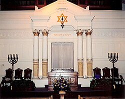 A white wall with an inset Torah ark at its center faces the viewer. Atop the ark is gold-colored Hebrew writing, and atop that a hanging lamp suspended from the center of a gold-colored Star of David. On each side of the ark are tall white columns, and beside them three ornate dark chairs and large seven branched menorahs. In front of the ark is a wooden pulpit.