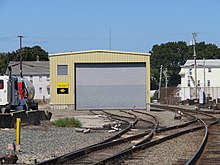 A small train yard is seen, with two of its tracks entering a large garage door. The building has a sign indicating it is owned by the Providence and Worcester Railroad. Another of the tracks continues past the building.