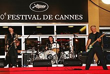 The Edge, Adam Clayton, and Larry Mullen Jr. performing outside a building on red carpet, with a sign in the background reading "Festival de Cannes".
