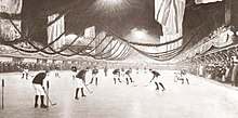 A group of hockey players are positioned on the ice inside the arena. The arena is decorated with flags on the sides. Surrounding the ice on all sides is a large group of spectators.