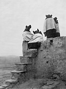 photogravure print of four young Hopi women in elaborate hairstyles with their backs to the camera, on the roof of an adobe building to better see dancers performing in the area below