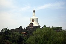 Color photograph of a white, bell-shaped building composed (from bottom to top) of a square base, three round disks of increasingly smaller diameter, a cut reverse cone, and a thinner tapering column with horizontal flutings crowned by the golden statue of a sitting figure. It appears to emerge from a forested area, against the background of a slightly cloudy blue sky.