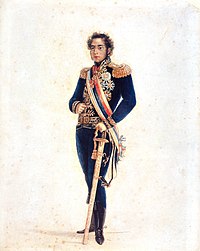 Colored full-length portrait showing a young man with curly hair and long sideburns who is wearing an elaborate gold-embroidered blue military tunic with gold epaulets and medals, blue trousers, black boots, a striped sash of office and gold belt, with his left hand resting on a sheathed sword