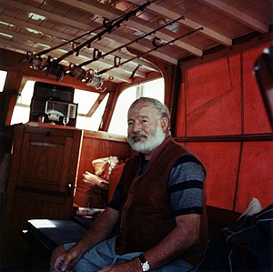 Photograph of an old bearded man sitting under fishing rods in a dimly lit ship's cabin.