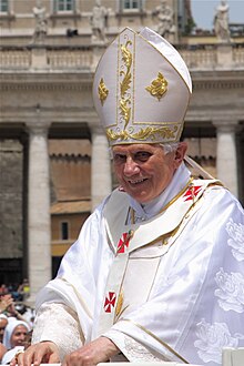 Farbfotografie vom Papst in liturgischer Kleidung und Papstkrone aus Stoff mit goldenen und roten Ornamenten. Im Hintergrund sind ein altes Gebäude und Menschen zu sehen.