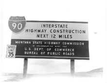 Black-and-white photograph of a sign with the I-90 shield announcing "Interstate Highway Construction Next 12 Miles" with a recommended speed limit of 35 miles per hour.
