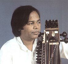 Ein Mann im mittleren Alter trägt ein Hemd und schaut zur Seite über ein Streichinstrument, das er nah am Körper hält.