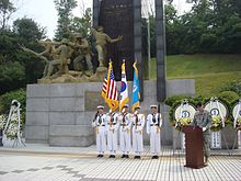 A line of sailors in white dress naval uniforms in front of a large monument while another soldier in green stands at a podium beside it