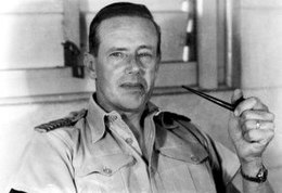 Informal portrait of man with short dark hair in light-coloured open-necked military shirt, holding a pipe