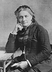 Retouched black-and-white photograph of an older woman, sitting next to a table on which she leans her right arm, with the hand supporting her head while her left hand holds a paper in her lap, dressed in black and with black lace covering most of her whitish hair